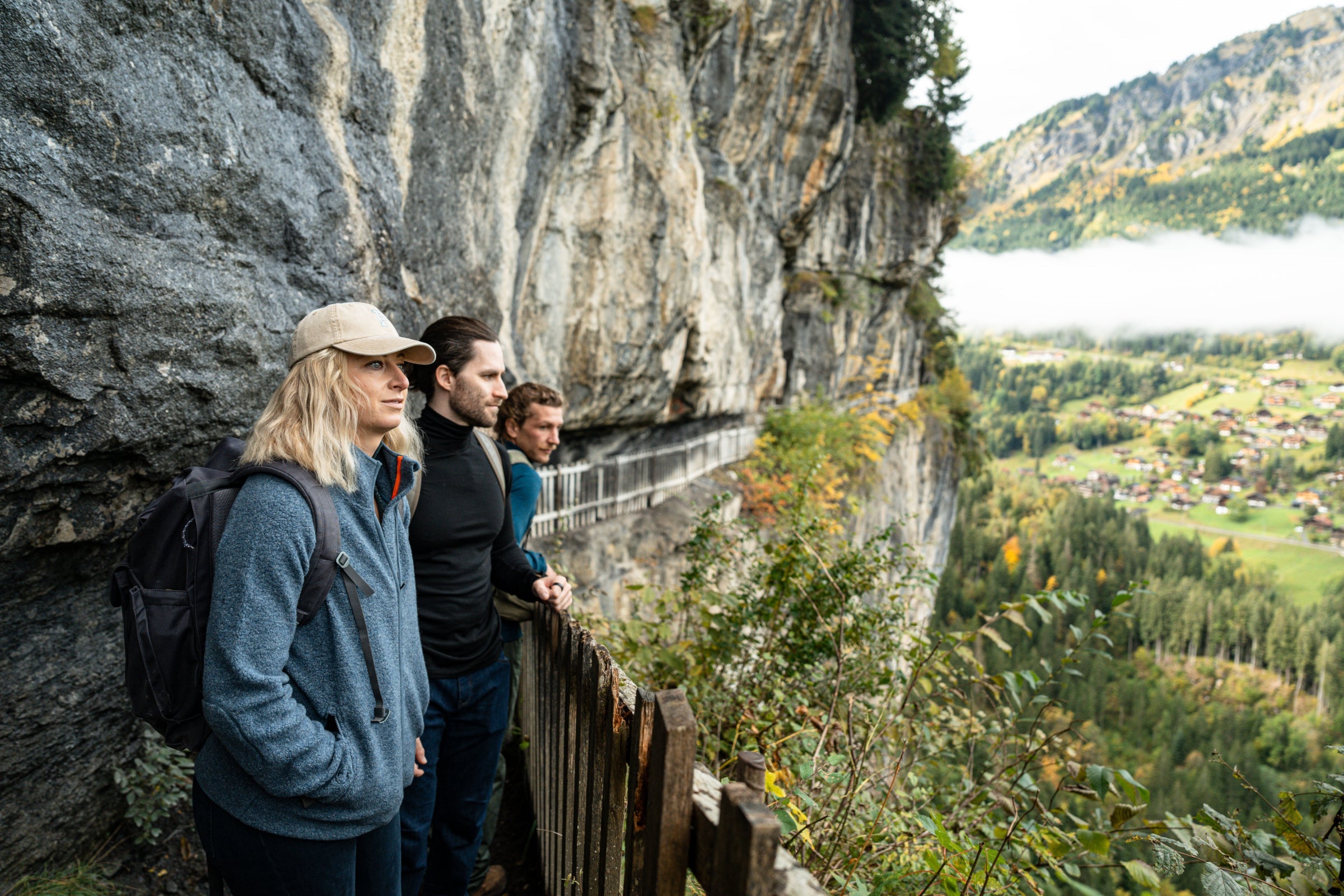 Three people standing on a cliffside path, overlooking a scenic valley with a village and forest below