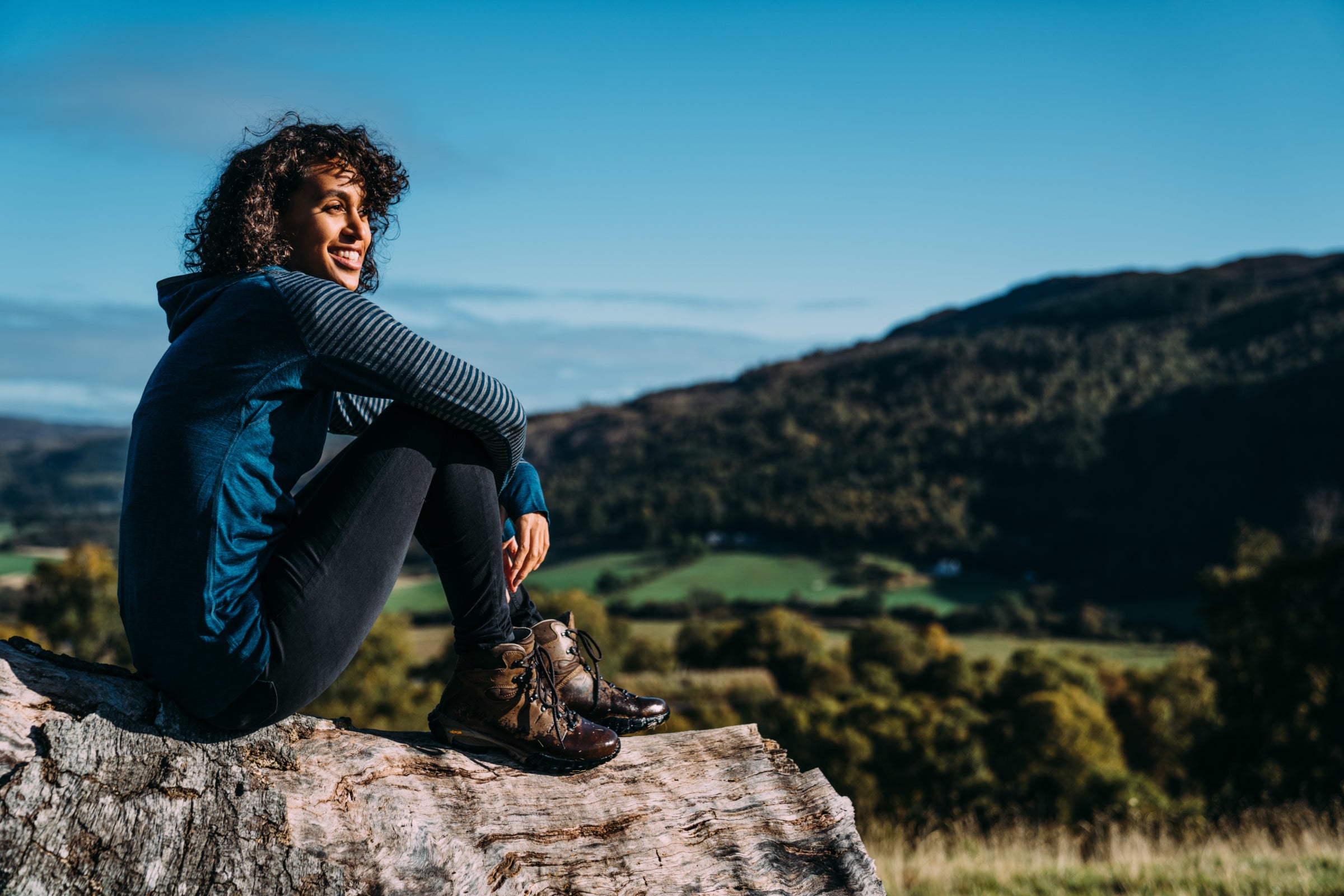 A person sits atop a log in the wilderness, dressed in adventure gear including a blue Isobaa merino wool zip top, smiling and enjoying the view