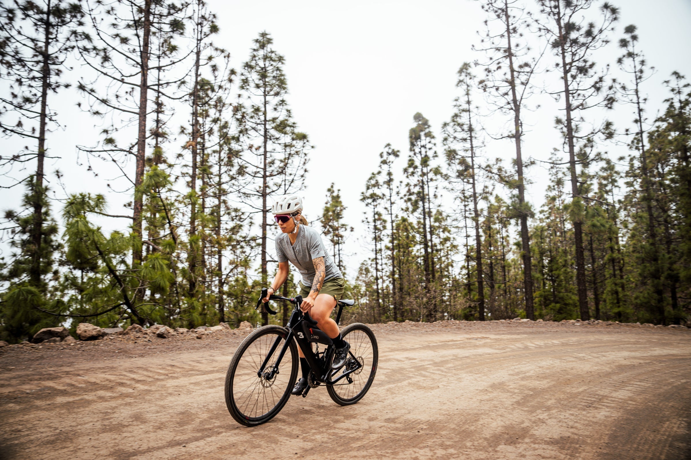 Woman cycling on a dirt road through a pine forest, wearing a Merino t-shirt and shorts