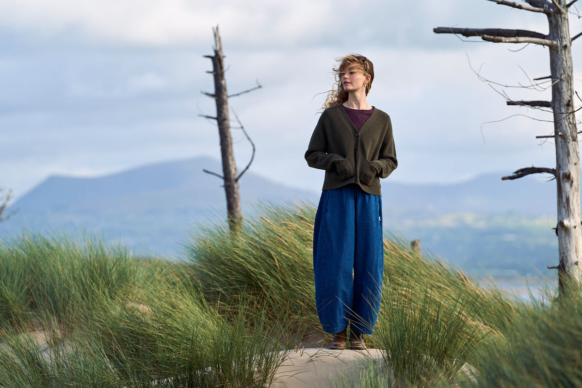 A woman stands outdoors on a stone path in a grassy field with mountains and cloudy sky in the background, wearing a dark brown cardigan over a burgundy top and loose blue trousers