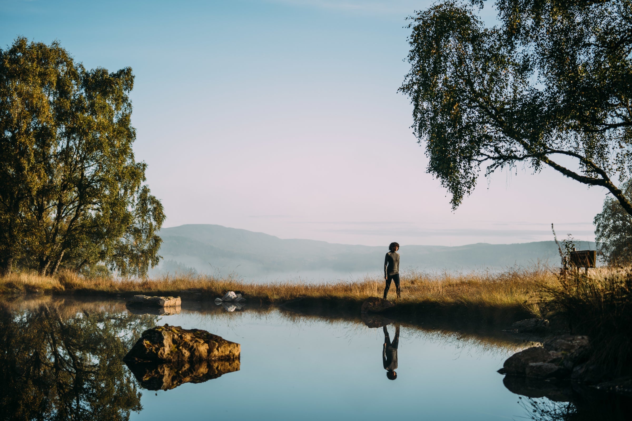 A person standing by a calm lake with trees and mountains in the background