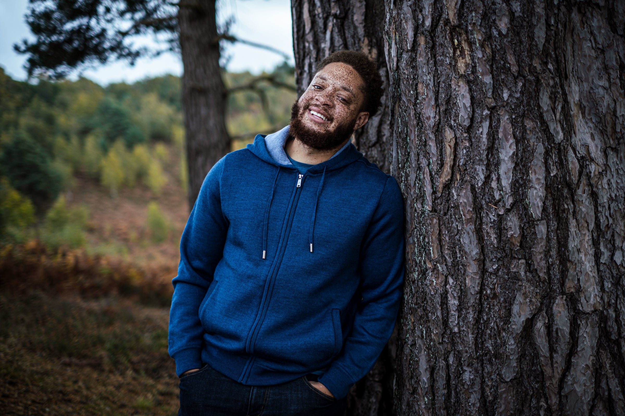 A man with a joyful expression, leaning against a tree in a forested area, wearing a blue Isobaa Merino Lux hoodie