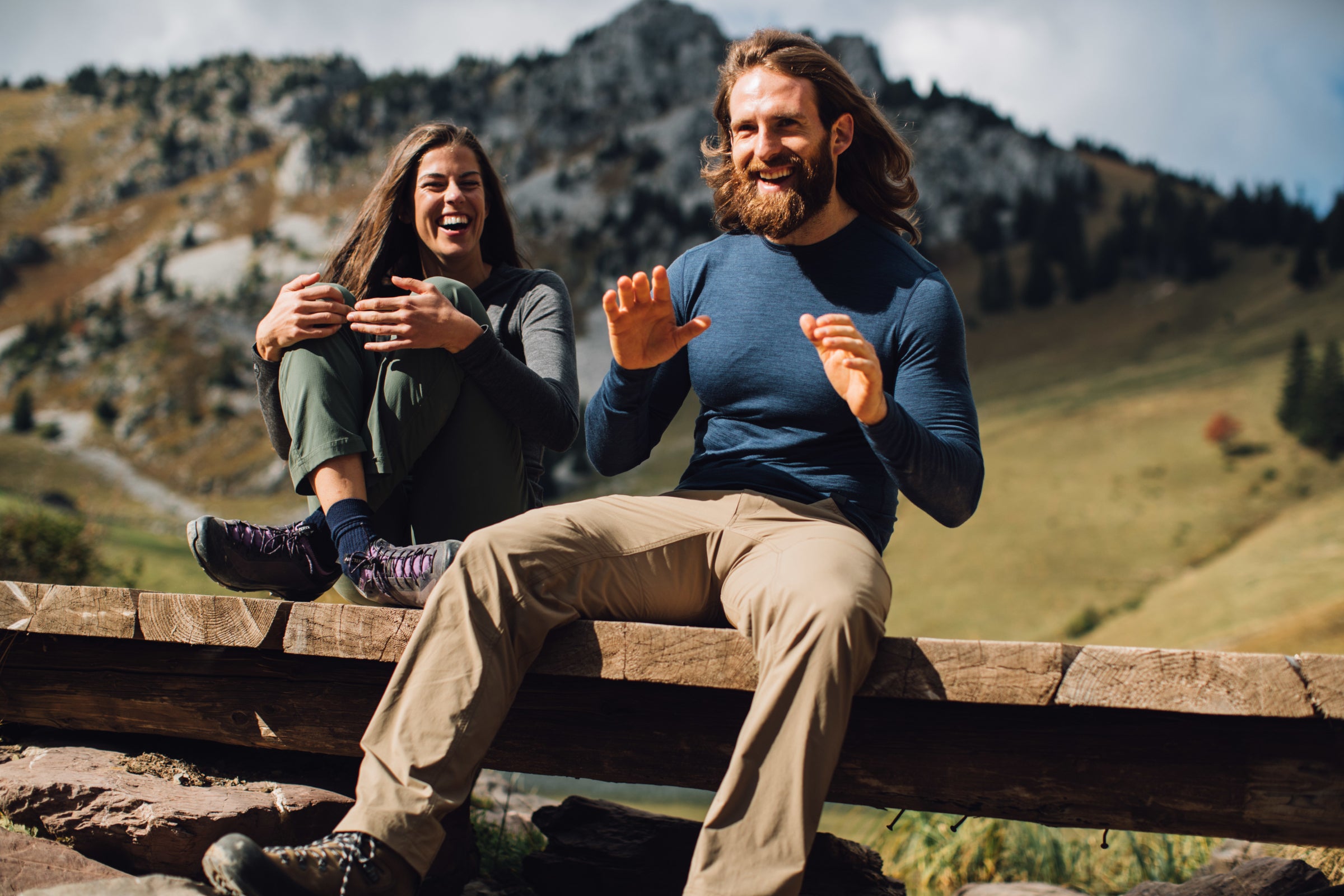 Two people sitting on a wooden bridge outdoors, laughing and enjoying the mountain scenery, both dressed in Isobaa Merino wool clothing