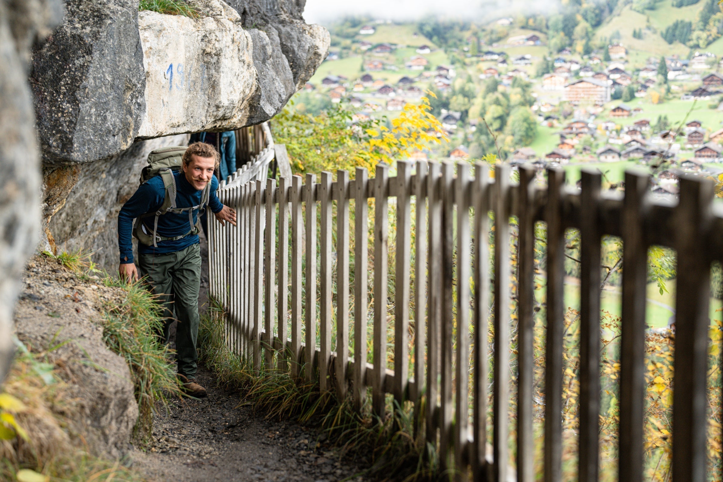 Hiker squeezing along a narrow cliffside trail with a wooden fence and valley below