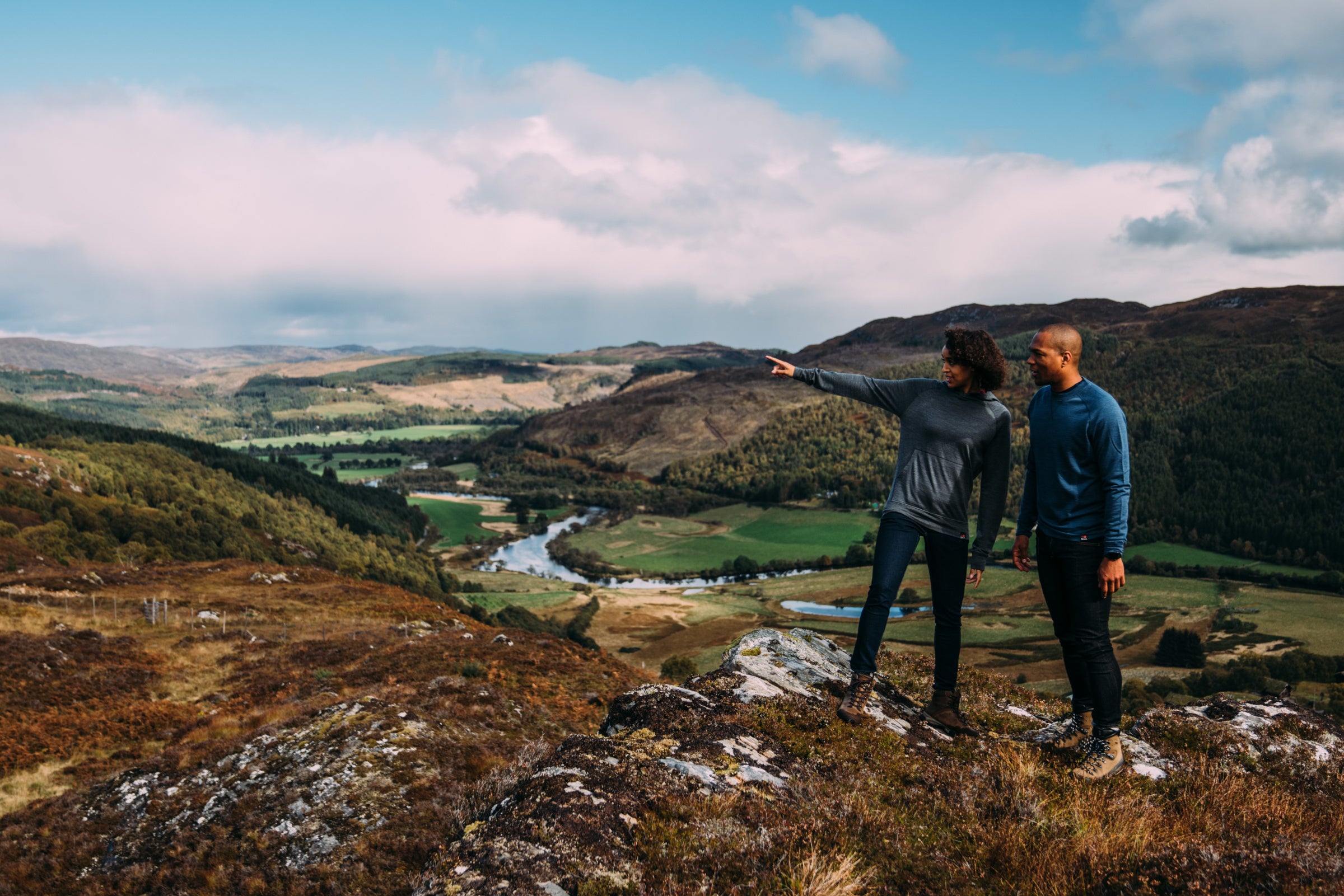 Two people standing on a rocky hilltop overlooking a scenic valley with a river. Both are dressed in casual Isobaa outdoor attire suitable for hiking. The landscape features rolling hills with patches of forest and open grasslands.