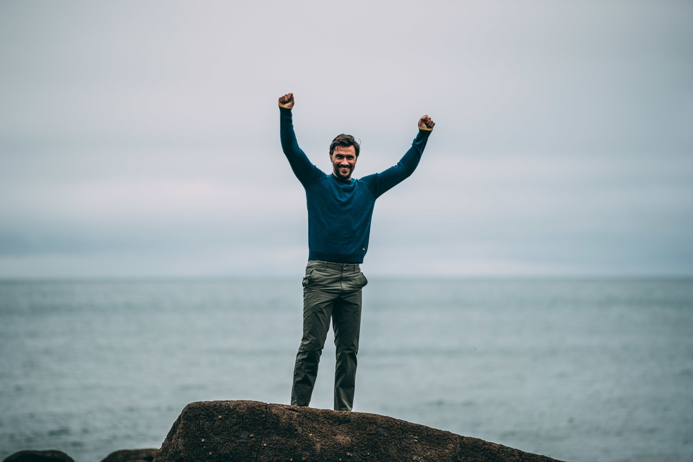 A man standing triumphantly on a rocky shore with his arms raised, wearing a blue merino sweater and green trousers, with an overcast sea in the background.