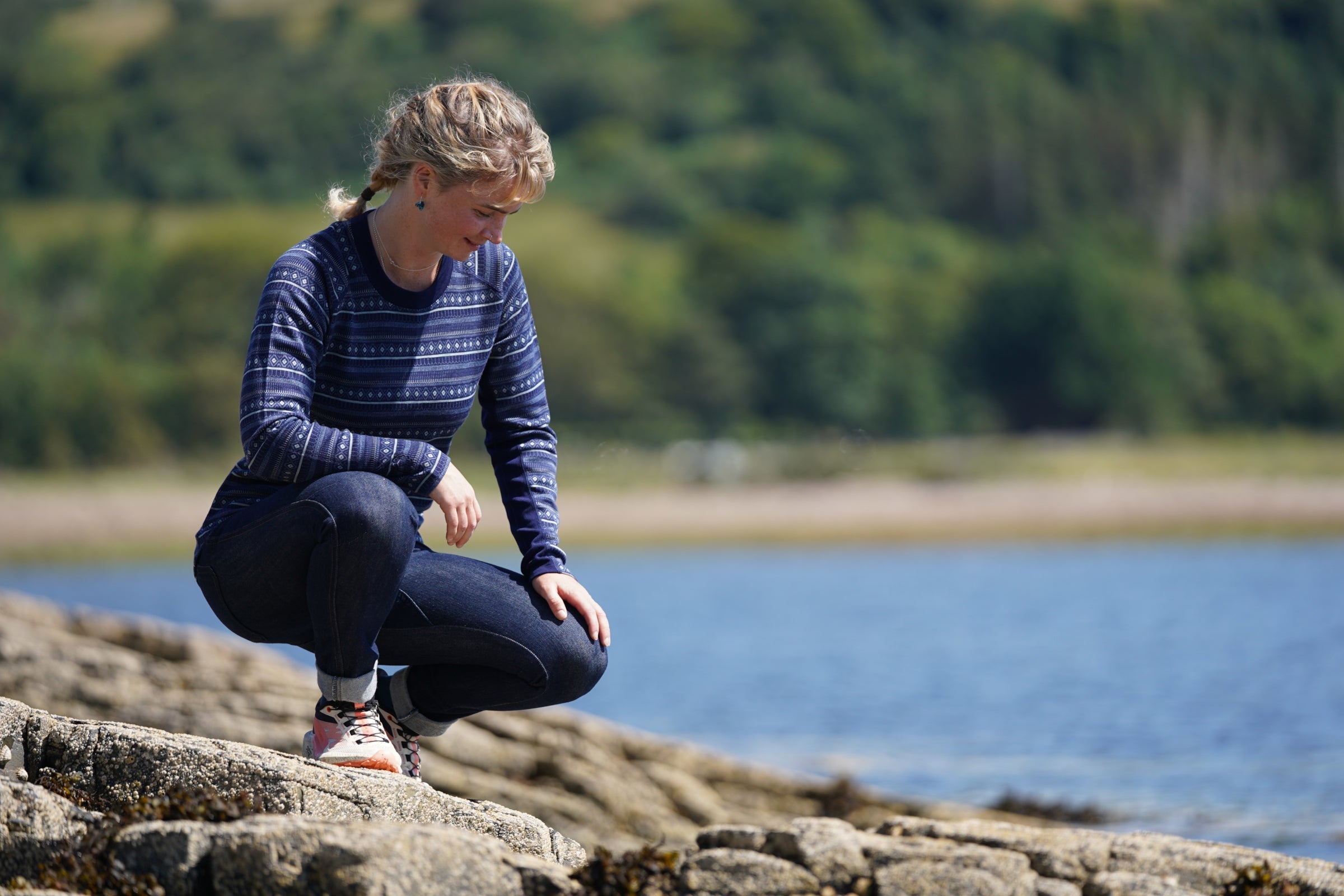 Person wearing a merino jacquard long-sleeve top crouching on rocks by the water