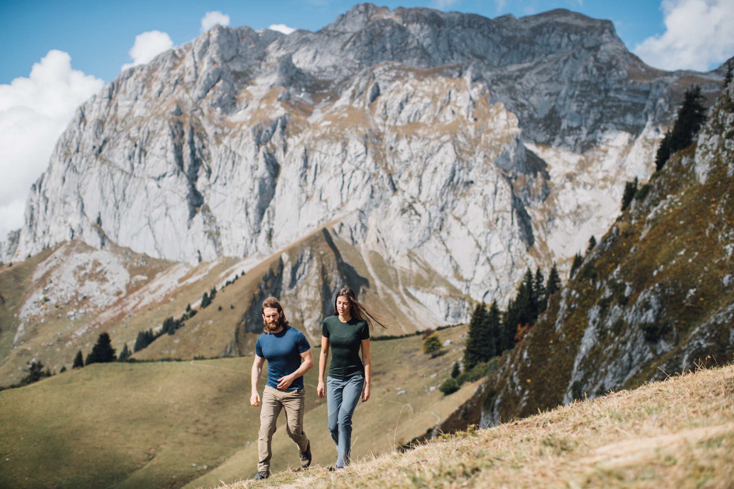 A man and a woman walking through a mountain meadow, both wearing Isobaa merino wool tops