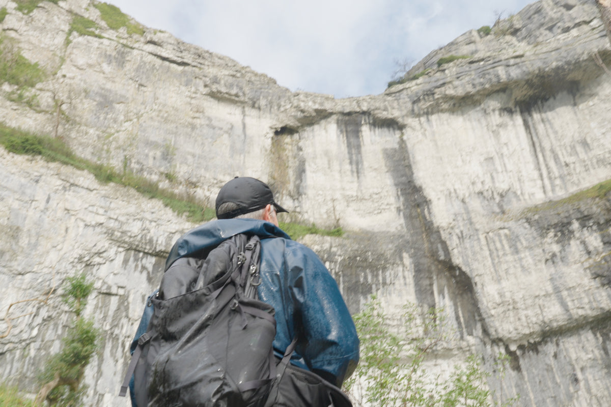 A person stands with their back to the camera, looking up at a massive pale rock cliff