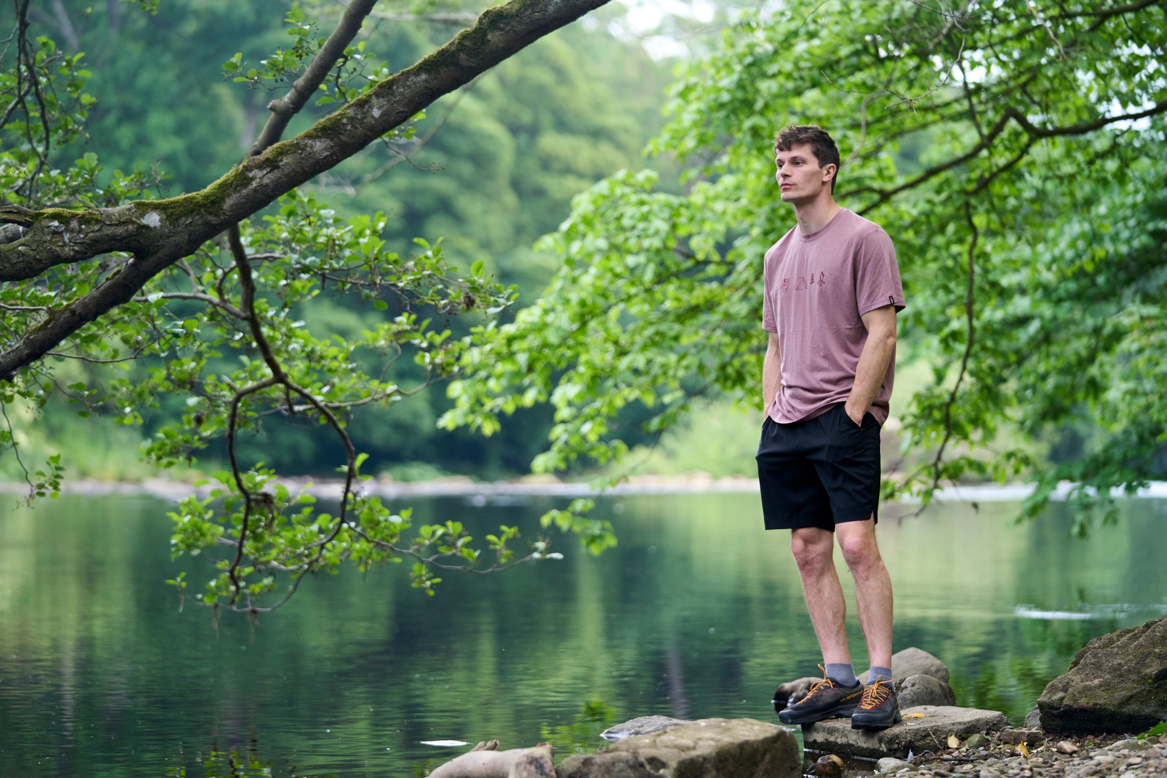 A man stands on rocks by a calm lake wearing a mauve-pink t-shirt