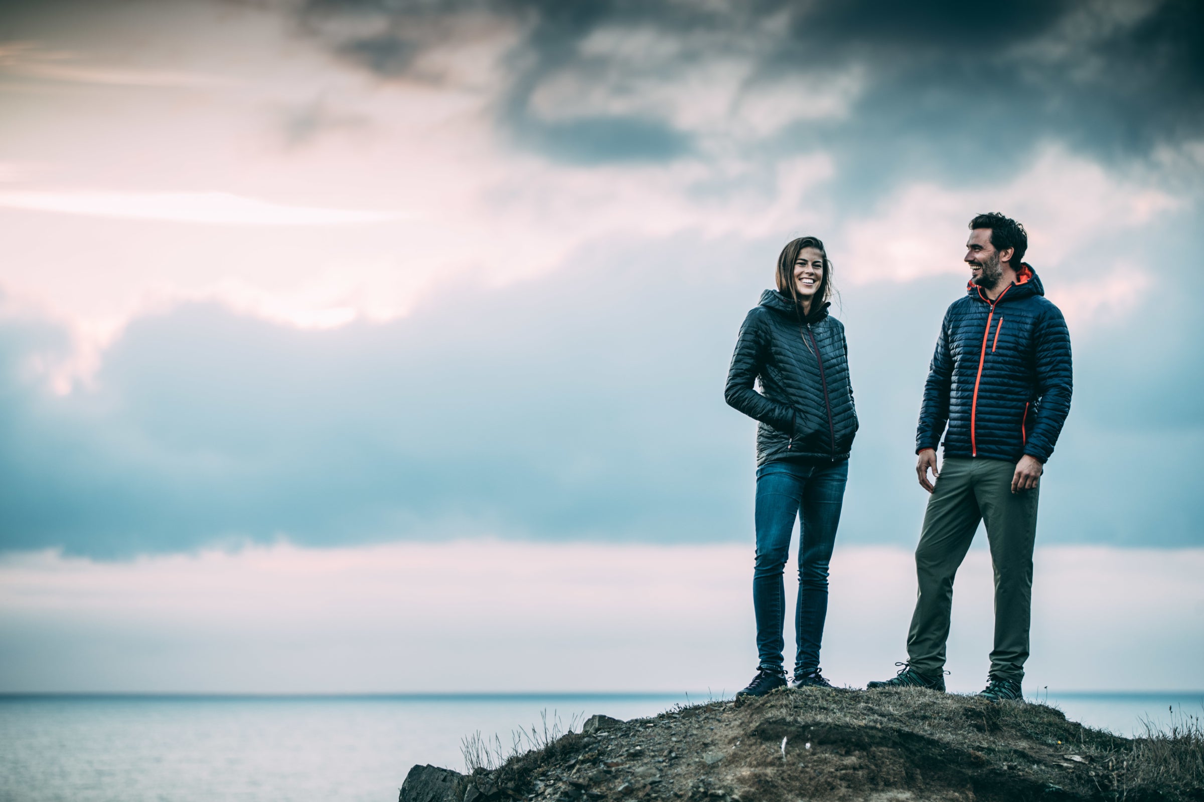 A man and a woman smiling at each other on a rocky hilltop, with a cloudy sky at sunset in the background, both wearing Isobaa merino insulated jackets