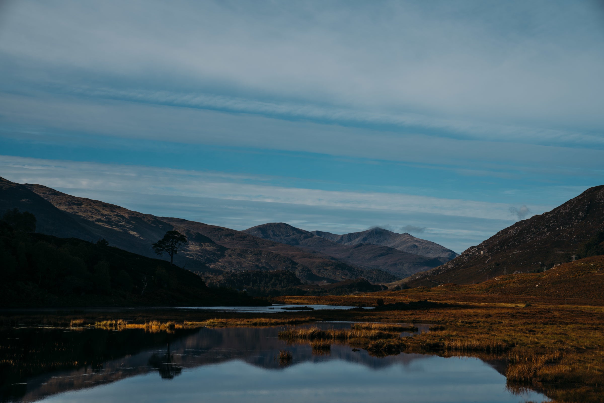 A calm lake reflecting mountains and blue skies with wispy clouds, surrounded by heather and grasses.