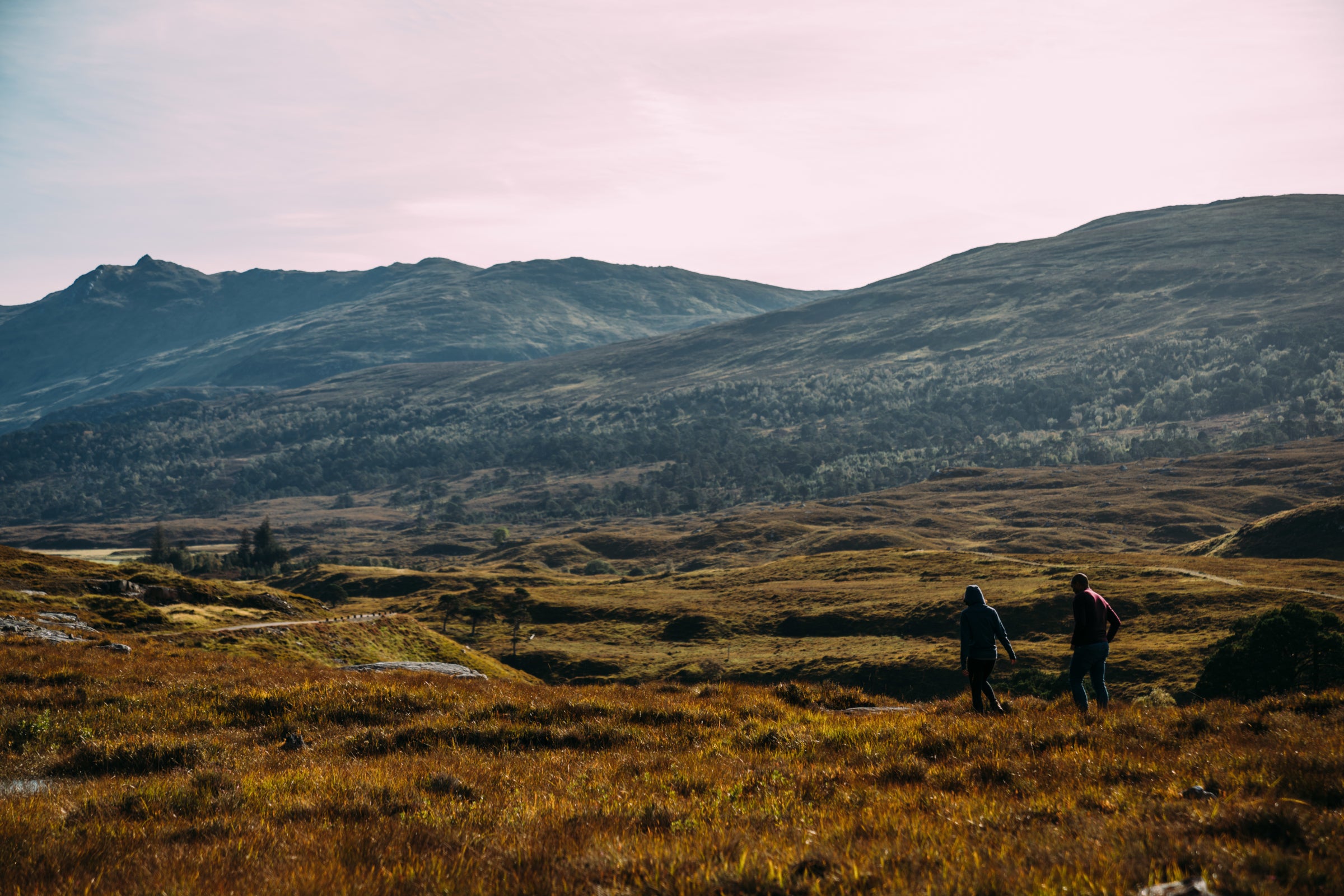 Two people hiking across a moor with rolling hills and mountain peaks in the distance under a warm sunlight.