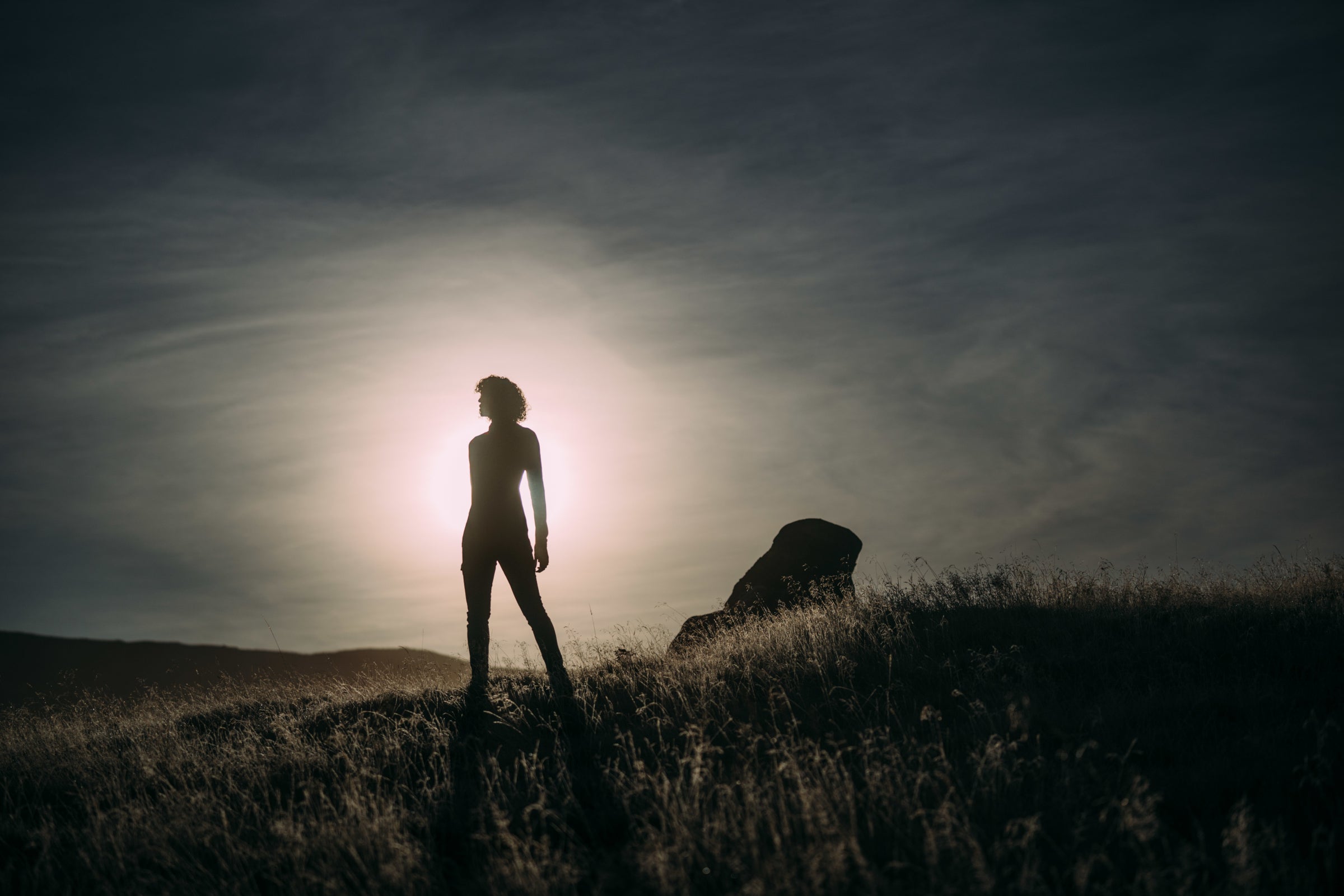 A silhouette of a person standing on a hilltop, backlit by the setting sun with a clear sky.