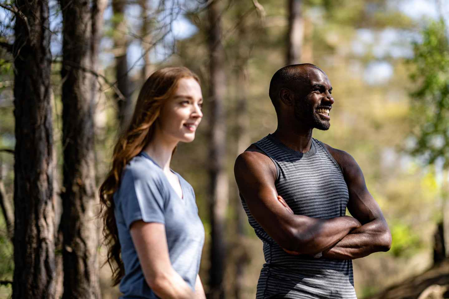 Man and woman outdoors in a wooded area, man in an Isobaa merino sleeveless top, woman in a light blue t-shirt