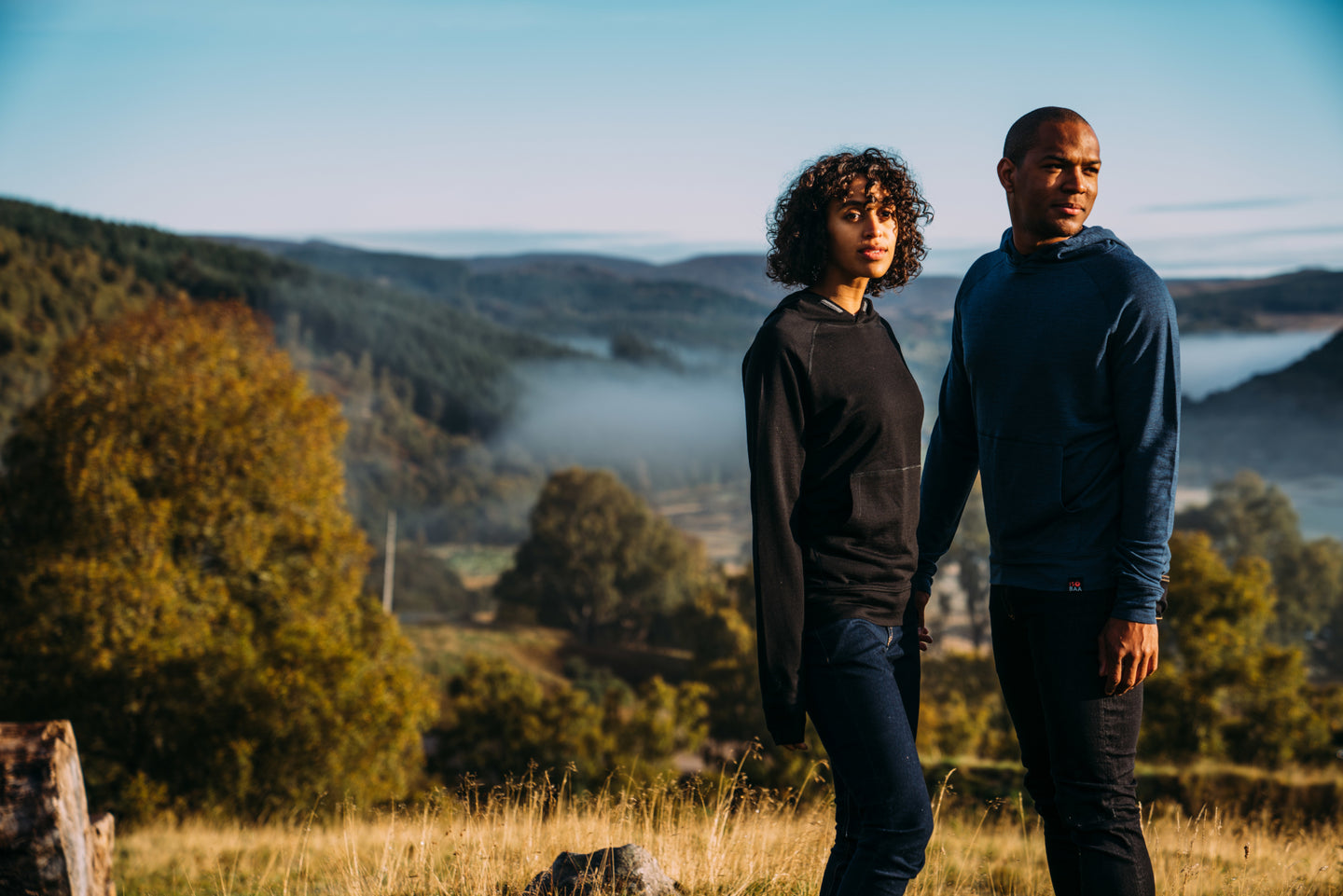 Man and woman standing in a scenic countryside, dressed in Isobaa merino hoodies