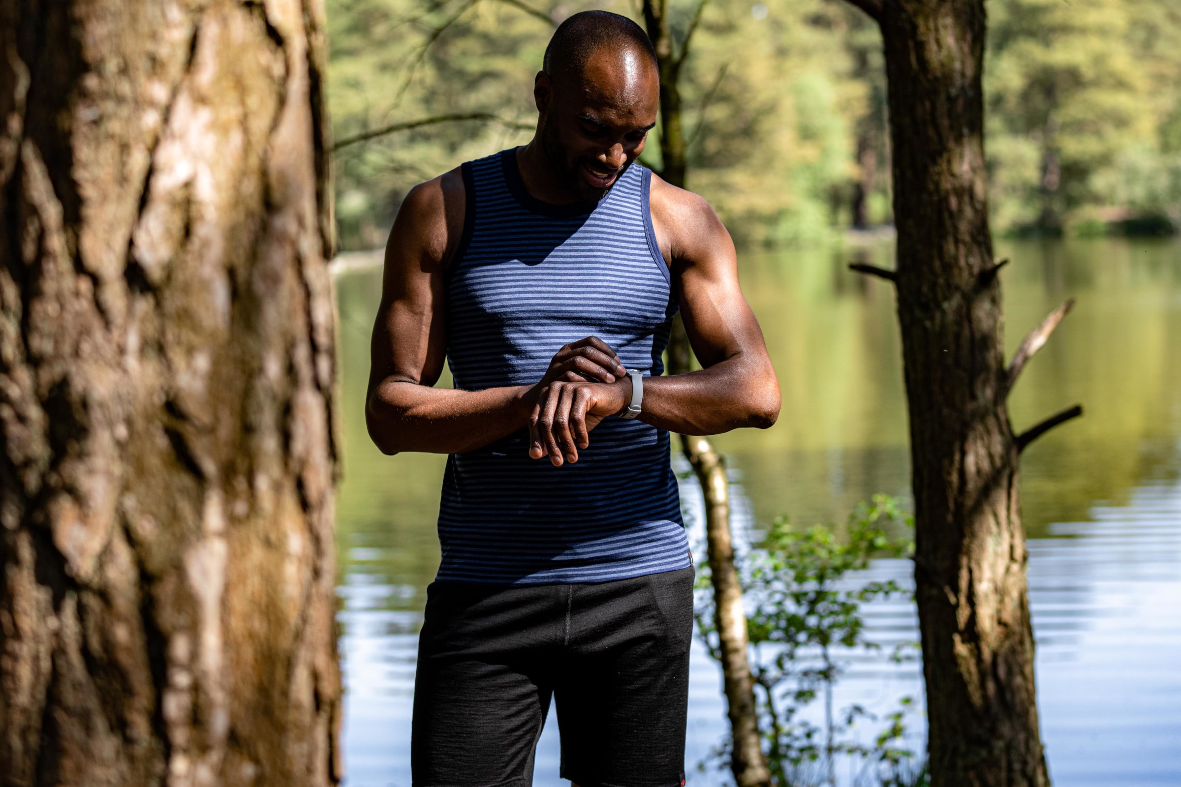 A man checking his fitness tracker, wearing a sleeveless merino top by Isobaa, standing near a lake