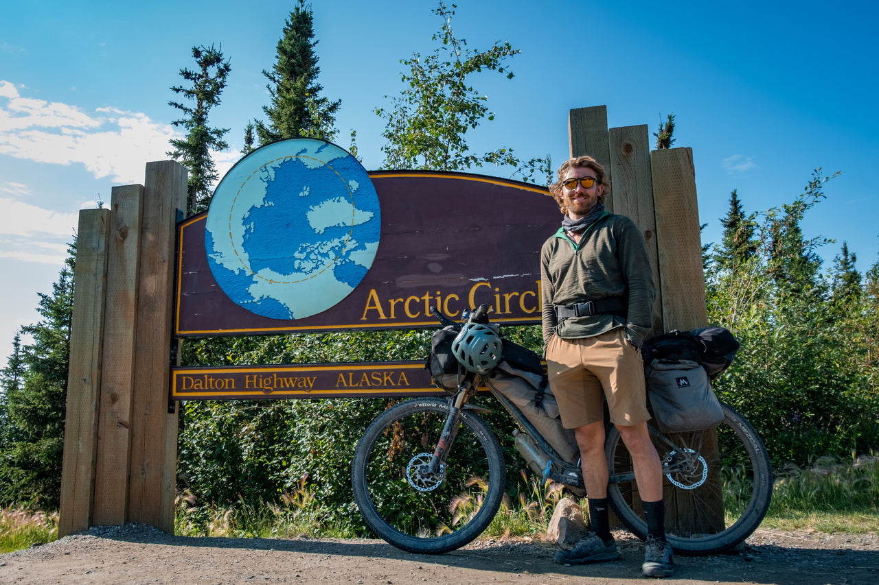 A bikepacker, and ambassador for Isobaa, Tristan Ridley, stands next to a bike, wearing Isobaa Merino wool clothing, at the Arctic Circle sign on the Dalton Highway in Alaska