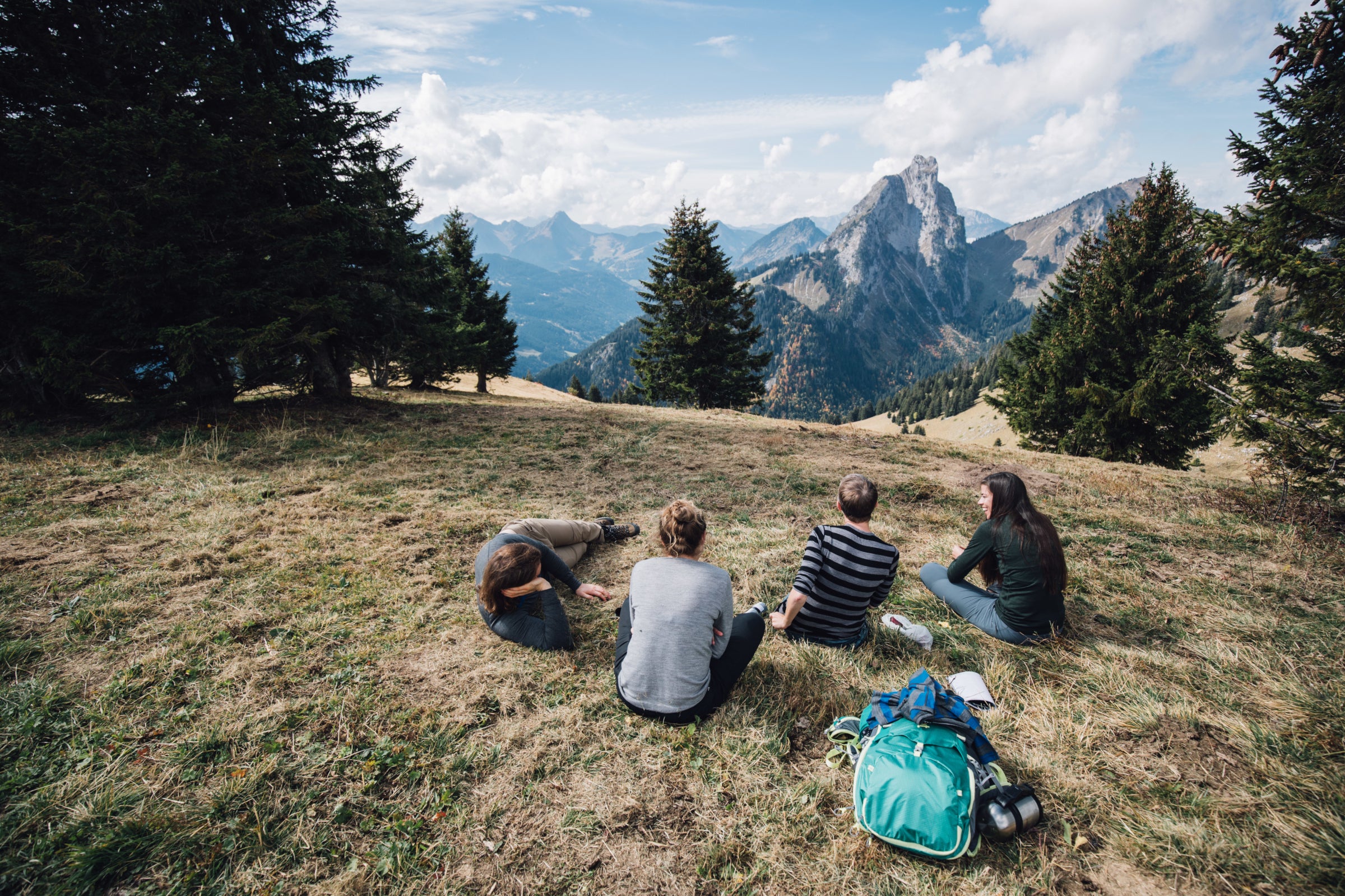 Four friends enjoying a serene mountain view, dressed in comfortable Isobaa merino wool clothing, with a backpack resting on the grass beside them