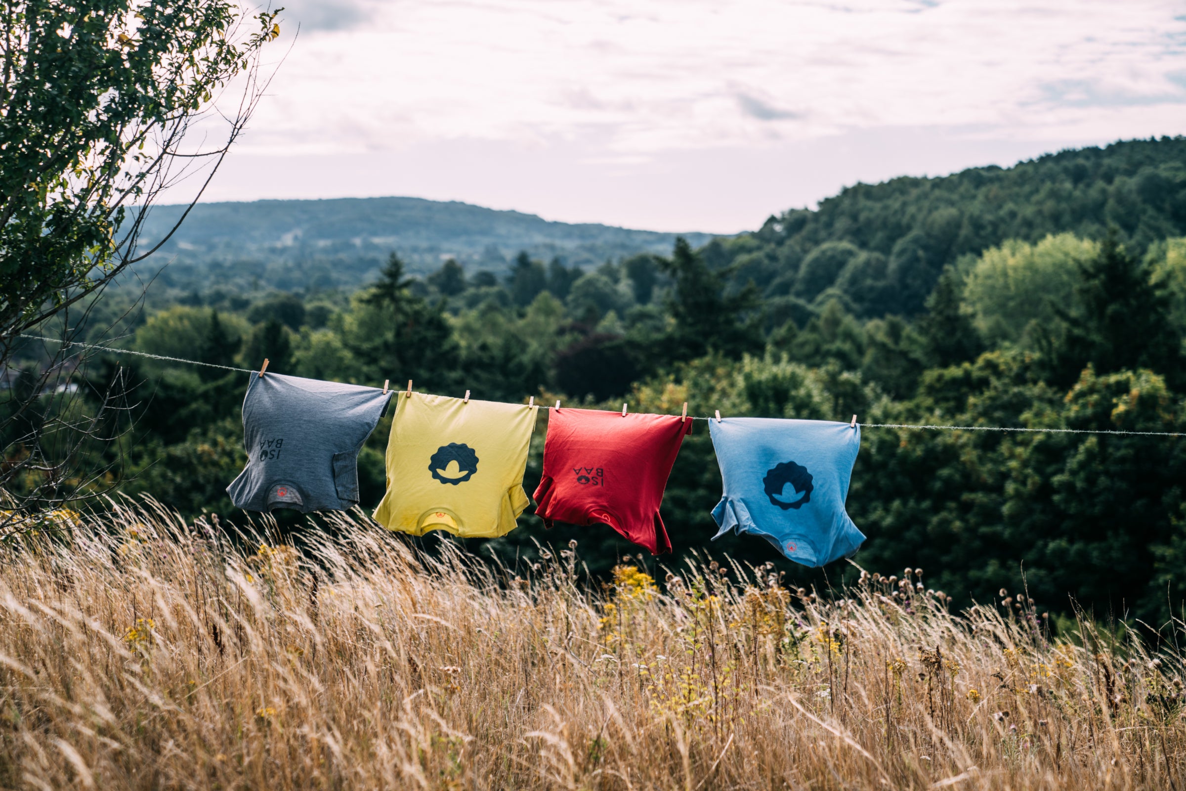 Four colorful Merino Wool T-shirts with printed Isobaa logos hanging on a clothesline in a sunny field with a backdrop of trees and hills.