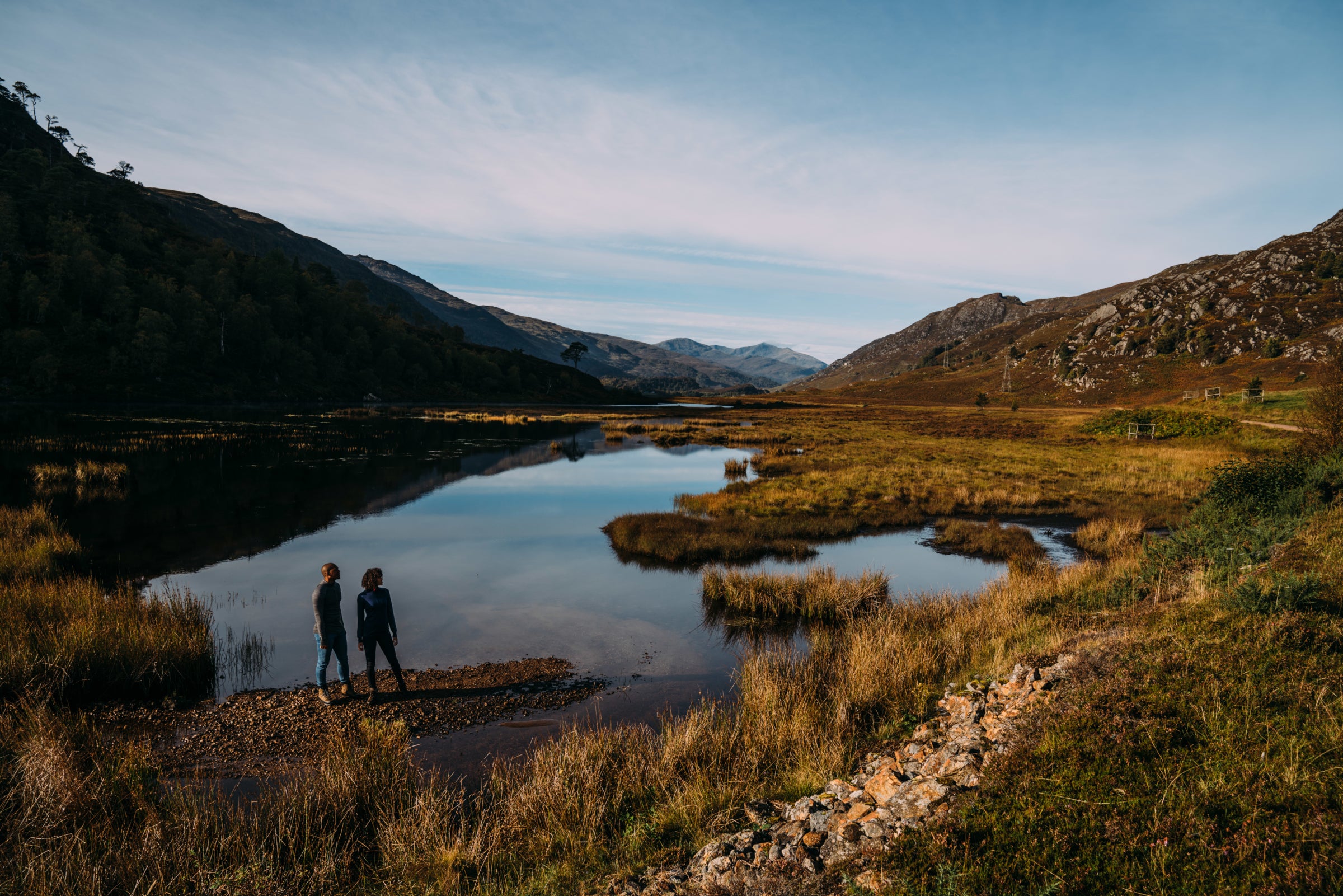 Two individuals stand by the serene waters of a mountain lake, surrounded by grassy plains and rolling hills under a soft pastel sky at dawn