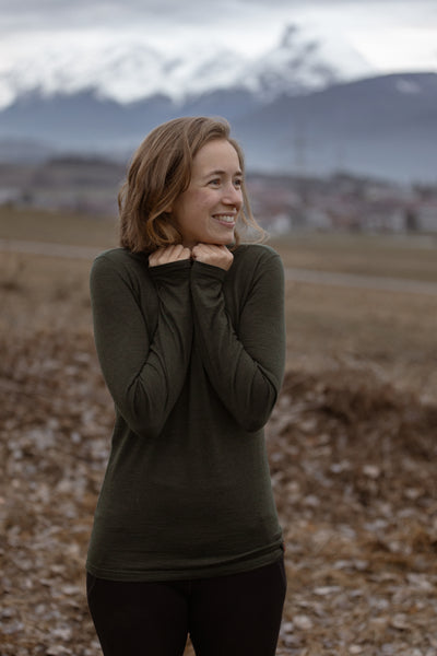 Smiling woman in green Isobaa merino top standing outdoors with misty mountains in the backdrop